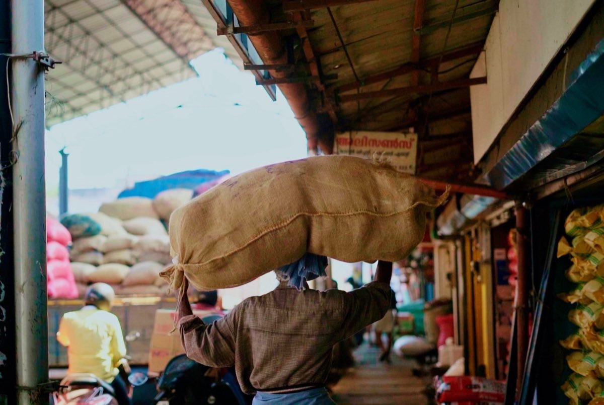 A worker carrying a jute sack through a wholesale market lined with stacked sacks